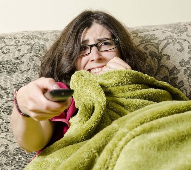 Woman with green blanket watches horror movies while alone on her couch.