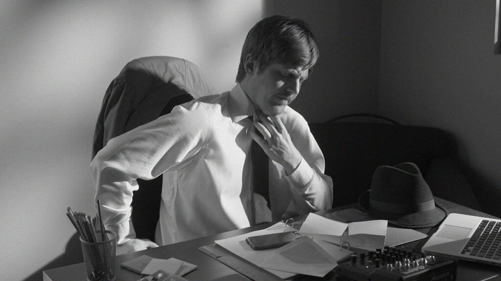 A man sits at a desk adjusting his tie in cinematic film noir style.