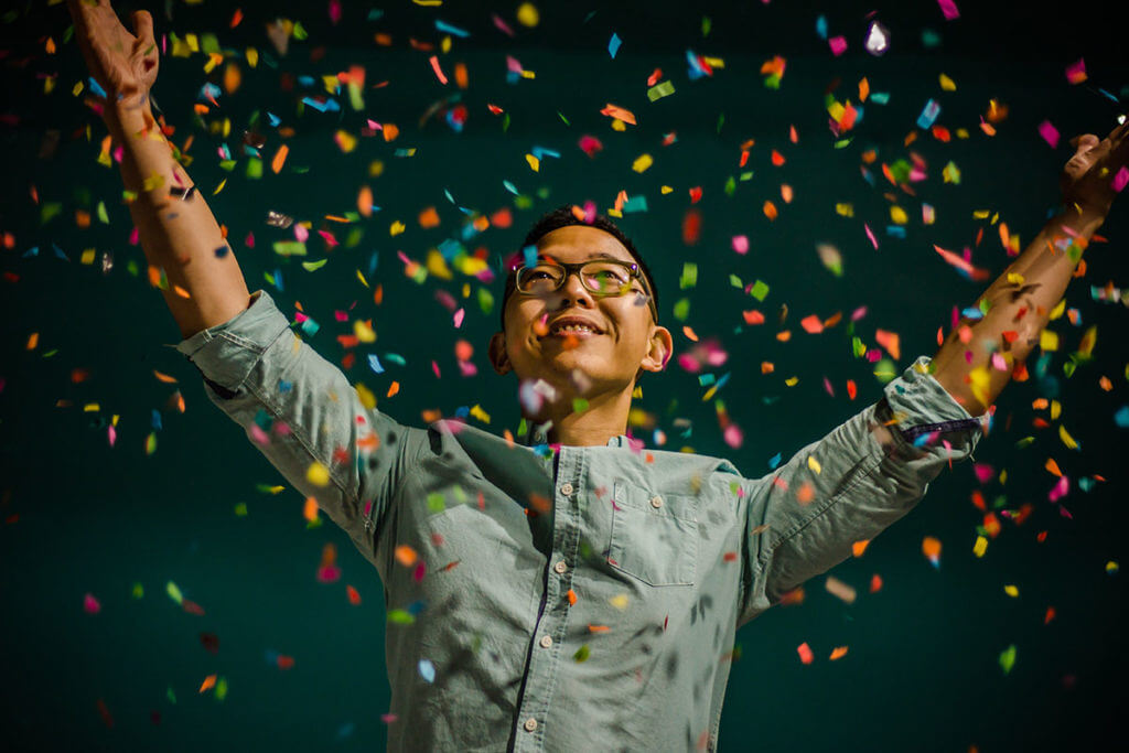 A confident man showered with confetti stands with arms raised after learning how to deal with imposter syndrome.