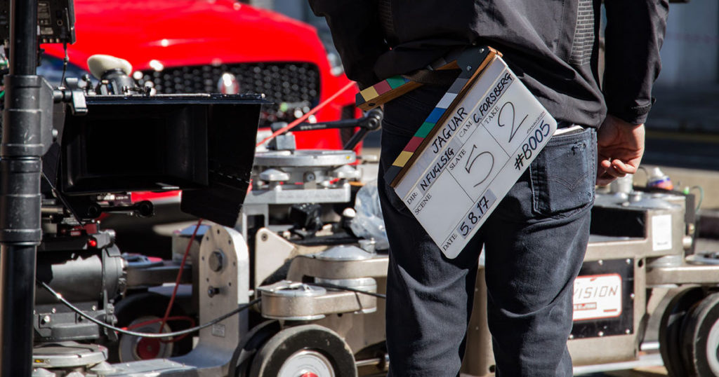 A person with a slate stands in front of equipment parked on set, indicative that locations cost money, baby!