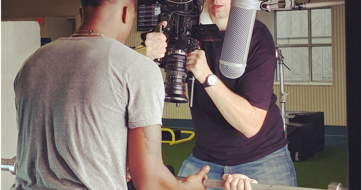 A man helps spot another man bench pressing while a camera operator films the weight lifter from above.