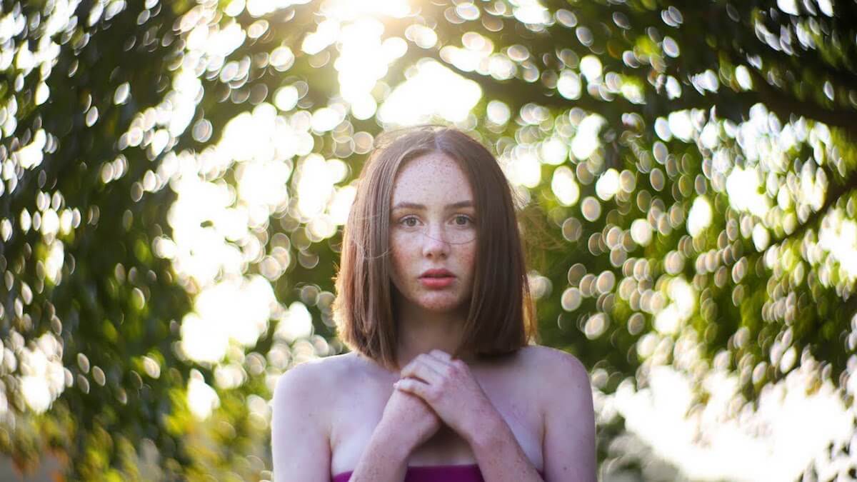 A young woman with trees behind her and her hands held to her chest stares into the camera for a bokeh portrait shot.