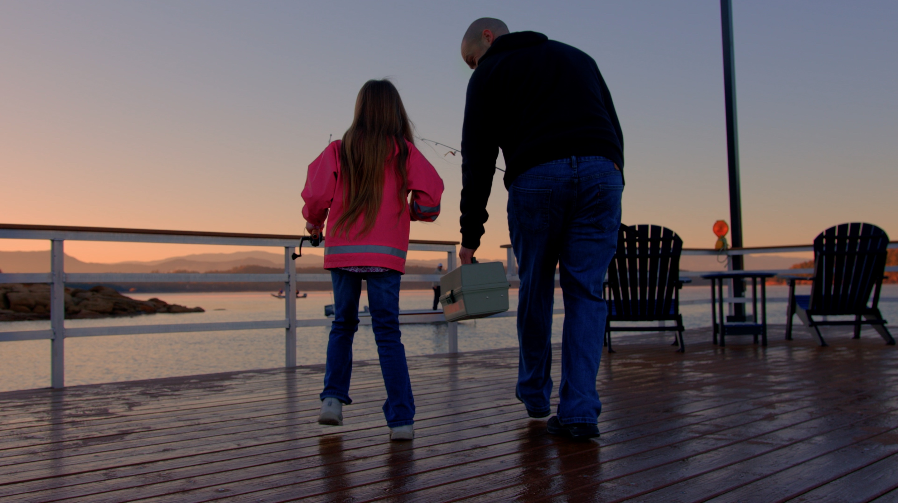 A man and little girl carry fishing equipment on a wet deck at sunrise.