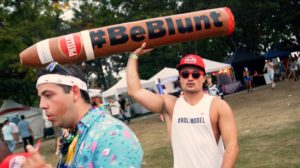 A man at a festival holding up a blunt that says #BeBlunt.