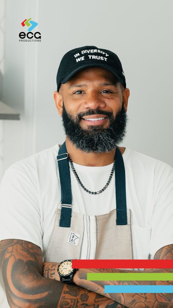 Justin Sutherland wearing a chef's coat and apron, confidently standing in a professional kitchen with culinary tools and plated dishes behind him—capturing his signature style and culinary expertise.