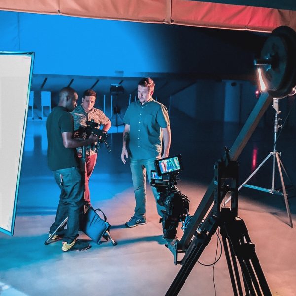 A jib in the foreground stands ready to film three men at an indoor shooting range.
