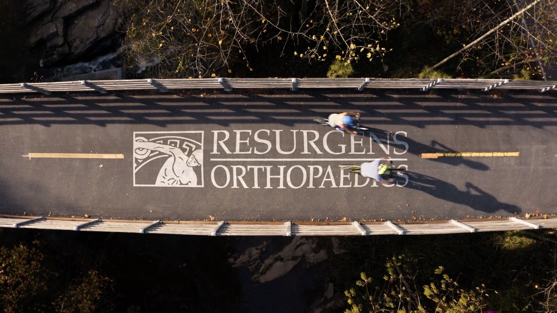 Two cyclists on the Silver Comet Trail pass over the words Resurgens Orthopaedics.