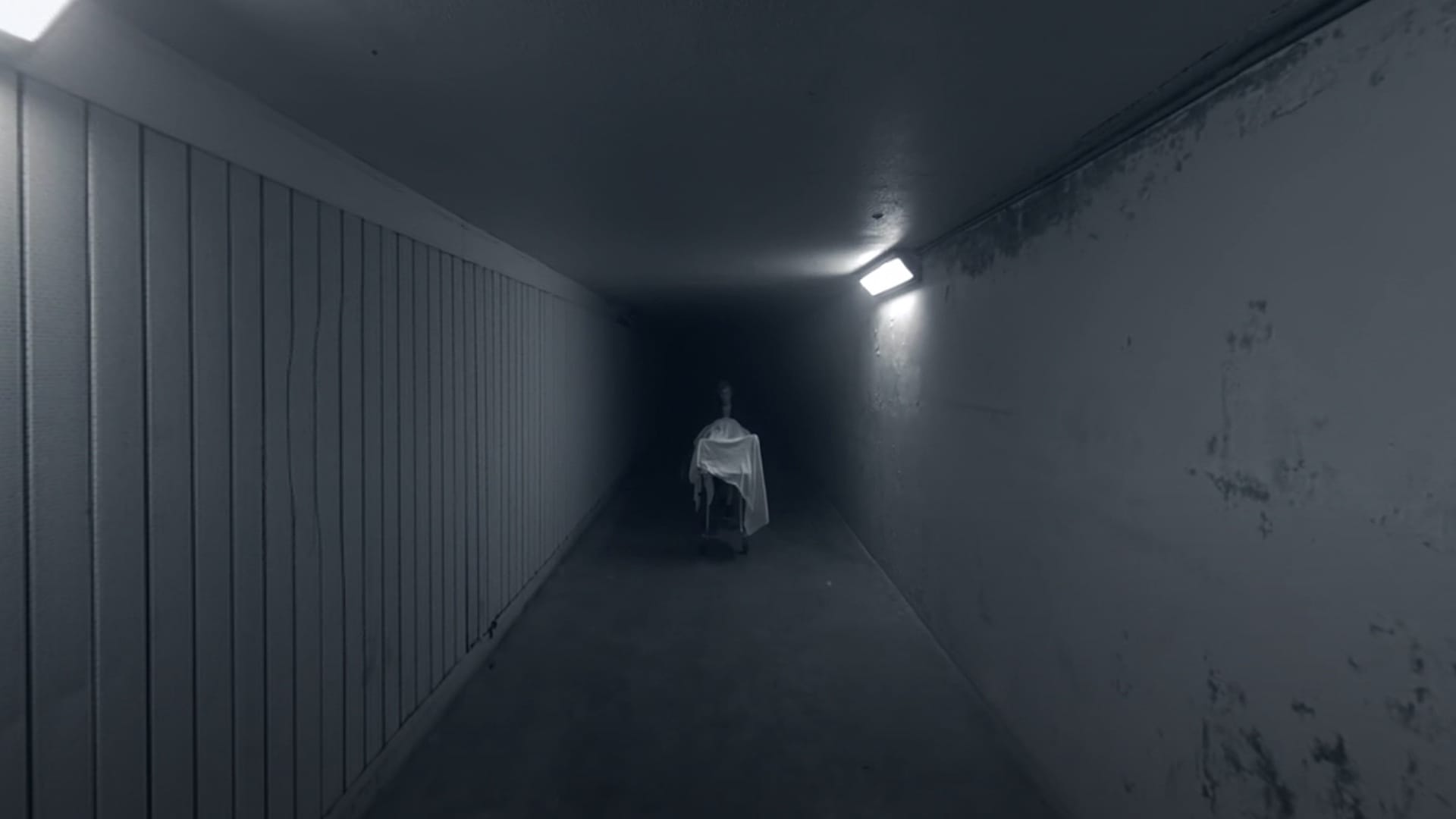 A cadaver covered by a sheet is wheeled down a dark hallway.